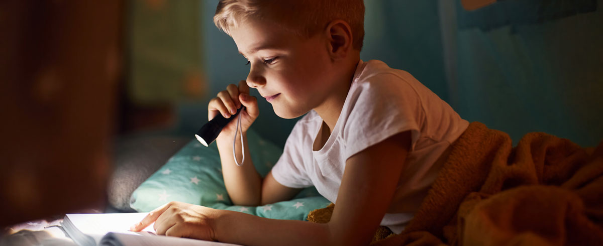 Een jongen die een boek leest met een zaklamp in zijn hand terwijl hij in bed ligt met een deken